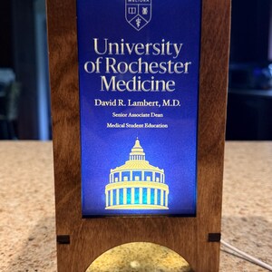 May include: A wooden framed plaque with the text "University of Rochester Medicine" on a blue background. The plaque also includes the name "David R. Lambert, M.D." and a building illustration. The frame is made of light brown wood.