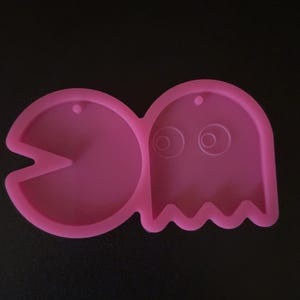 Puede incluir: Un molde de silicona rosa con Pac-Man y un personaje fantasma. La forma de Pac-Man tiene la boca abierta, mientras que el fantasma tiene ojos y un borde inferior ondulado. El molde está diseñado para manualidades o la creación de objetos decorativos.