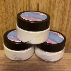 May include: Three small, white jars with black lids are stacked on a wooden surface. Each jar has a label with text and a blue and pink design. The jars contain a white, creamy substance.