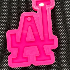 May include: A bright pink silicone mold in the shape of the Los Angeles Dodgers logo, featuring the letters "LA". The mold is designed for crafting and creating custom items. The background is a black surface with white dots.