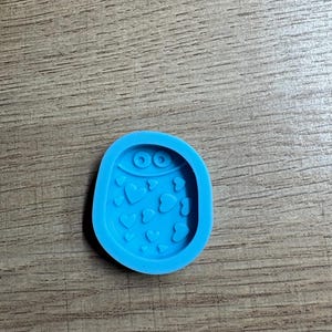 May include: A light blue silicone mold in an oval shape, featuring a design of hearts and two eye-like circles at the top. The mold is designed for crafting and creating decorative elements.