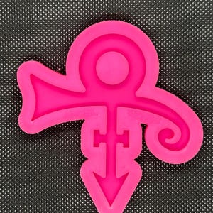 May include: A pink silicone mold shaped like the iconic Prince symbol. The symbol is a combination of a stylized dove, a cross, and an arrow.