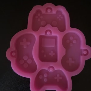 Puede incluir: Molde de silicona rosa con varios diseños de mandos de videojuegos, incluyendo una consola portátil y varios estilos de gamepad. El molde está diseñado para la elaboración y creación de artículos temáticos de juegos.