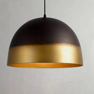 K&ouml;nnte beinhalten: Eine Pendelleuchte mit einem halbkugelf&ouml;rmigen Schirm. Die obere H&auml;lfte ist dunkelbraun, die untere H&auml;lfte goldfarben. Das Licht h&auml;ngt an einem schwarzen Kabel und erzeugt ein modernes, minimalistisches Design.