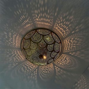 Pode incluir: Uma luminária de teto decorativa redonda, de cor bronze, com um padrão de pequenos círculos. A luz projeta sombras intrincadas na superfície circundante, criando um jogo de luz e sombra.