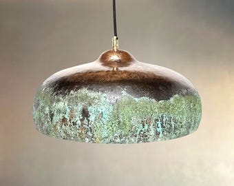 May include: A dome-shaped pendant light fixture with a copper finish. The top is a dark brown color, transitioning to a green and turquoise patina on the lower half. The light hangs from a black cord with a brass-colored fitting.