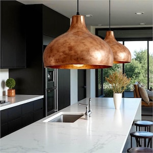 May include: Two copper-colored pendant lights hang above a white countertop with a stainless steel sink. The kitchen features black cabinets and a view of the outdoors through a large window. A vase of dried flowers sits on the counter.