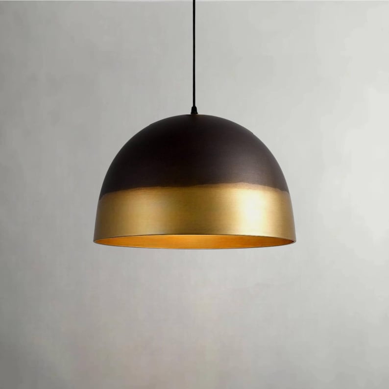 K&ouml;nnte beinhalten: Eine Pendelleuchte mit einem halbkugelf&ouml;rmigen Schirm. Die obere H&auml;lfte ist dunkelbraun, die untere H&auml;lfte goldfarben. Das Licht h&auml;ngt an einem schwarzen Kabel und erzeugt ein modernes, minimalistisches Design.