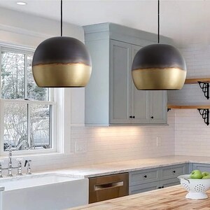 Puede incluir: Dos lámparas colgantes modernas con un diseño bicolor. La mitad superior de cada lámpara es gris oscuro, mientras que la mitad inferior es dorada. Las lámparas cuelgan de cables negros en una cocina.