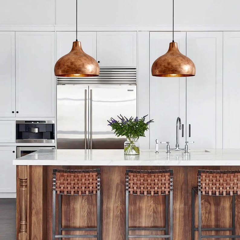 May include: Two copper-colored pendant lights hang above a kitchen island with woven bar stools. The kitchen features white cabinets, a stainless steel refrigerator, and a marble countertop. A vase of purple flowers sits on the counter.