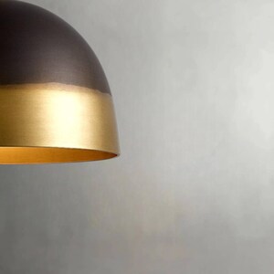 K&ouml;nnte beinhalten: Eine moderne Pendelleuchte mit zweifarbigem Design. Der obere Teil ist dunkelbronze, w&auml;hrend der untere Bereich eine goldene Oberfl&auml;che aufweist. Die Leuchte hat eine abgerundete, kuppelf&ouml;rmige Form, und der Innenraum ist ebenfalls goldfarben.