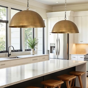 May include: Two large, dome-shaped pendant lights with a brushed brass finish hang from the ceiling. Each fixture features a wide, rounded metal shade suspended by a thin chain, providing downward illumination over a kitchen island area.