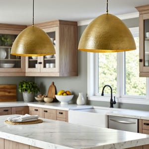 May include: Two gold-coloured pendant lights hang above a marble countertop in a kitchen. The lights have a textured surface and a dome shape. The kitchen features wooden cabinets, a white sink, and a window.