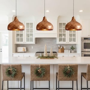 May include: Three copper-colored pendant lights hang above a white kitchen island. The lights have a bell shape with a textured finish. Wicker bar stools with green wreaths are in front of the island. The kitchen has white cabinets and a white countertop.