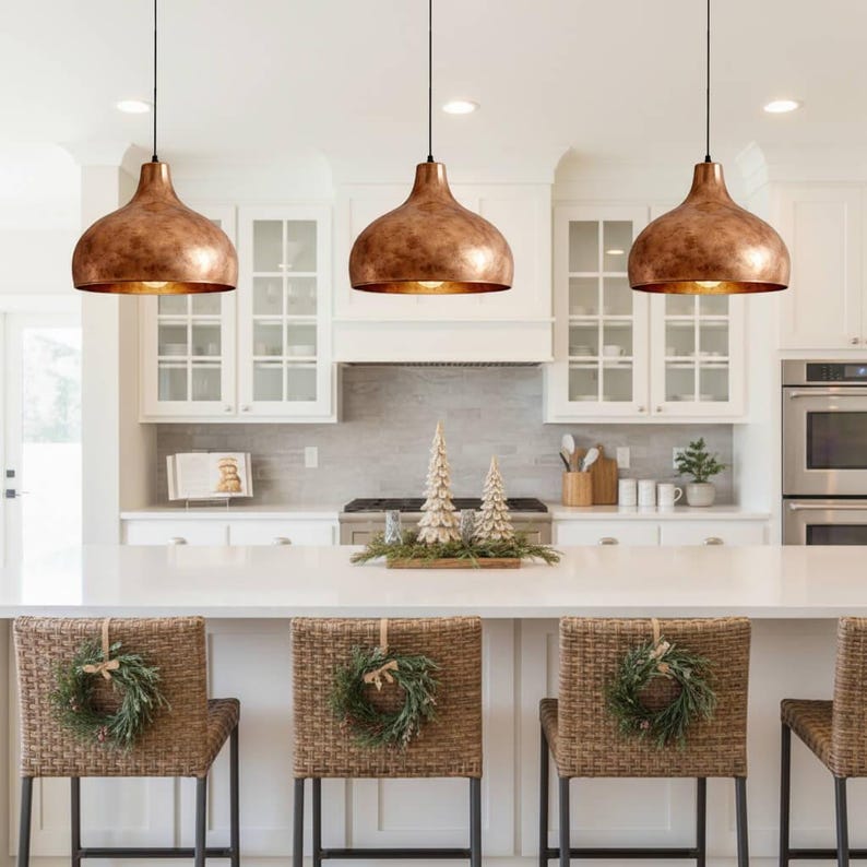 May include: Three copper-colored pendant lights hang above a white kitchen island. The lights have a bell shape with a textured finish. Wicker bar stools with green wreaths are in front of the island. The kitchen has white cabinets and a white countertop.