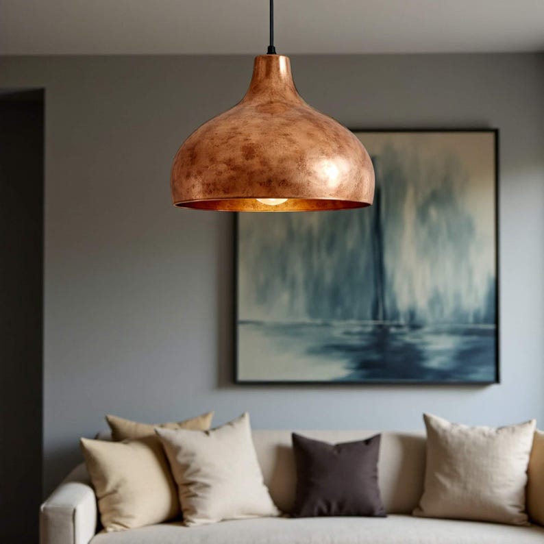 May include: A copper-colored pendant light fixture with a bell-shaped shade hangs from a black cord. The light is suspended above a cream-colored sofa with several pillows. A painting with blue and white tones is visible in the background.