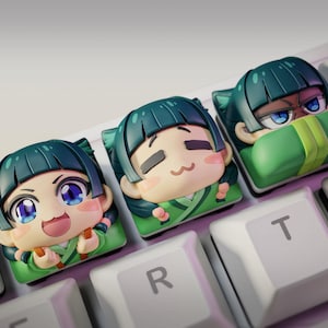 Könnte beinhalten: Drei Keycaps im Anime-Stil mit unterschiedlichen Gesichtsausdrücken, die jeweils einen Charakter mit teal-farbenem Haar und einem grünen Outfit zeigen. Die Keycaps befinden sich auf einer weißen Tastatur, mit dem Logo "nom nom figures" oben links.