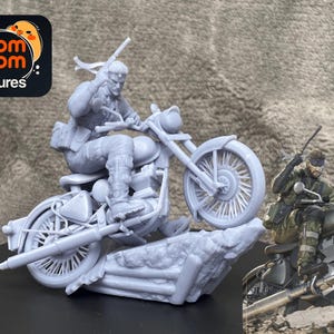 Puede incluir: Una figura gris de un personaje montando una motocicleta, con una segunda figura de un personaje en una motocicleta en el fondo. La figura está sobre una base rocosa. El logotipo "nom nom Figures" está en la esquina superior izquierda.