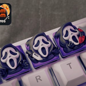 Può includere: Tre tasti personalizzati con la maschera di Ghostface della serie di film horror Scream. Ogni tasto ha una maschera bianca, un cappuccio nero e una base viola. Uno tiene un coltello, un altro un telefono.