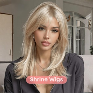Puede incluir: Una persona con cabello rubio largo y en capas y flequillo. Lleva una sudadera con capucha gris oscuro. El texto "Shrine Wigs" se muestra en una pancarta rosa en la parte inferior de la imagen.