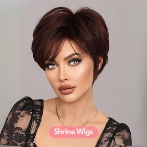 Puede incluir: Peluca corta color castaño rojizo con flequillo lateral. La peluca está peinada con volumen en la coronilla y la lleva una persona de piel clara, ojos azules y un collar dorado. El texto "Shrine Wigs" es visible.