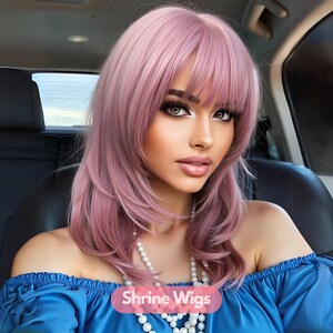 Può includere: Una donna con lunghi capelli rosa con frangia, indossa una blusa blu senza spalle e una collana di perle. Il testo "Shrine Wigs" è visibile nell'immagine.