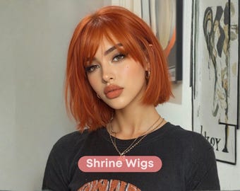 May include: A woman with orange-red hair and a black t-shirt. The hair is styled in a short bob with bangs. The t-shirt has the text "Shrine Wigs" on a pink oval. The woman is wearing gold jewelry.