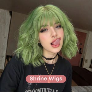 Puede incluir: Una persona con cabello verde ondulado y flequillo, que lleva una camiseta negra y un collar. La imagen incluye el texto "Shrine Wigs" en un rectángulo rosa. La persona tiene un piercing en la nariz y saca la lengua.
