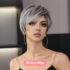 May include: A woman with a short, layered, silver and gray wig. She is wearing gold hoop earrings and a gold necklace. The text "Shrine Wigs" is displayed on a pink banner.