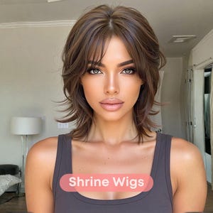 Op de afbeelding: Een vrouw met bruin haar en een gelaagde coupe. Ze draagt een grijze tanktop. De tekst "Shrine Wigs" staat op de voorkant van de tanktop. De achtergrond is een kamer met een lamp en een bank.