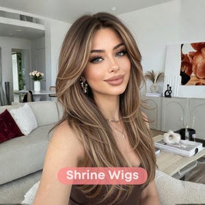May include: A woman with long, layered brown hair with blonde highlights. She is wearing a brown top and jewelry. The text "Shrine Wigs" is displayed in a pink oval at the bottom of the image. The background includes a sofa, artwork, and decorative items.