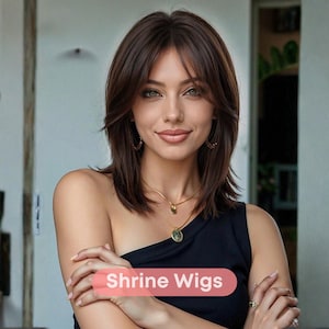 May include: A woman with brown hair and green eyes is wearing a black top and gold jewelry. The image includes the text "Shrine Wigs" in a pink banner. The woman is smiling and looking at the camera.