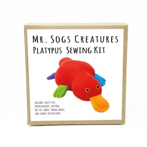 May include: A brown box with the text "MR. SOGS CREATURES PLATYPUS SEWING KIT" on the front. The box contains a red, green, blue, and yellow felt platypus sewing kit. The kit includes safety eyes, hypoallergenic stuffing, pre-cut fabric, thread, a needle, and simple instructions.