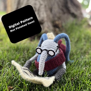 May include: A crocheted gray and blue creature with glasses is reading a newspaper while sitting in the grass. The text on the black square reads "Digital Pattern Not Finished Item!"