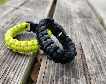 Può includere: Due braccialetti in paracord, uno nero e uno giallo e verde. Entrambi i braccialetti hanno una chiusura in plastica nera. I braccialetti sono su una superficie di legno invecchiato.