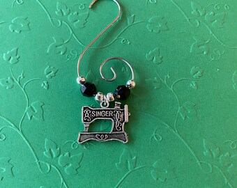 May include: A silver sewing machine charm with the word "SINGER" is suspended from a silver hook. Two black faceted beads and two silver beads are above the charm. The background is a green surface with a floral pattern.