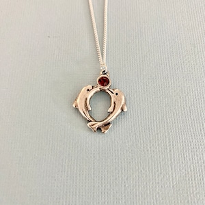 May include: Silver necklace with a pendant of two dolphins intertwined in a heart shape with a red gemstone in the centre.