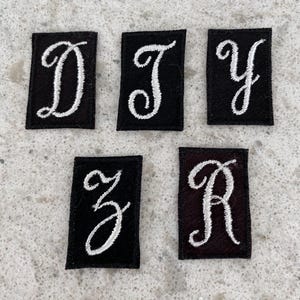 May include: Five black felt patches with white embroidered letters. The letters are D, J, Y, Z, and A. The letters are in a cursive font.