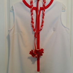 May include: A white sleeveless dress with a red trim and a red beaded necklace with pink accents. A red fabric flower is attached to the front of the dress.