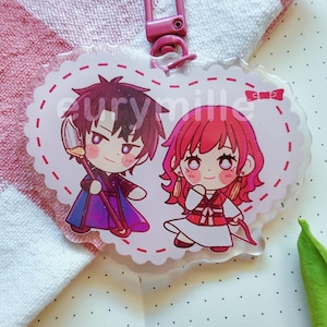 May include: A heart-shaped acrylic keychain featuring a chibi-style illustration of two characters. One character has dark hair and a purple outfit, while the other has pink hair and a red and white outfit. The text eurymille is printed in white.