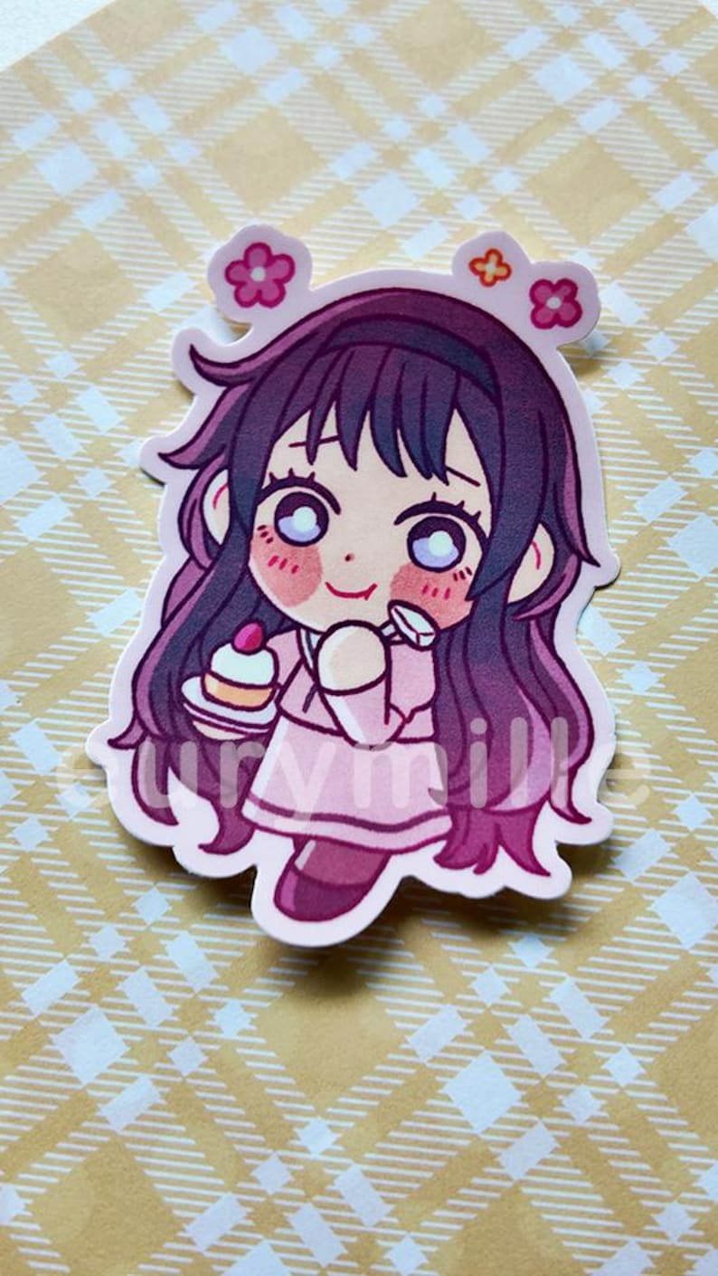 May include: A pink sticker featuring a cartoon girl with long, dark purple hair and large eyes. She wears a pink dress and holds a small cake. The sticker has a white border and flower accents.