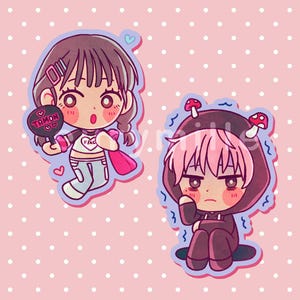 以下が含まれることがあります： アニメキャラクターのちびキャラステッカー2枚。一人は茶髪でハート付きの白いトップスを着て、TAMONと書かれた黒い扇子を持っている。もう一人はピンクの髪で、2つの赤いキノコが付いた暗い色のフード付きの服を着ている。