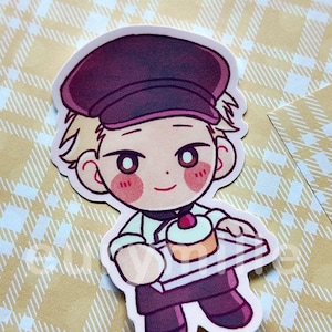 May include: A die-cut sticker featuring a cartoon character in a baker's uniform, holding a tray with a cupcake. The character has blonde hair, a maroon hat, and rosy cheeks. The background is a yellow plaid pattern.