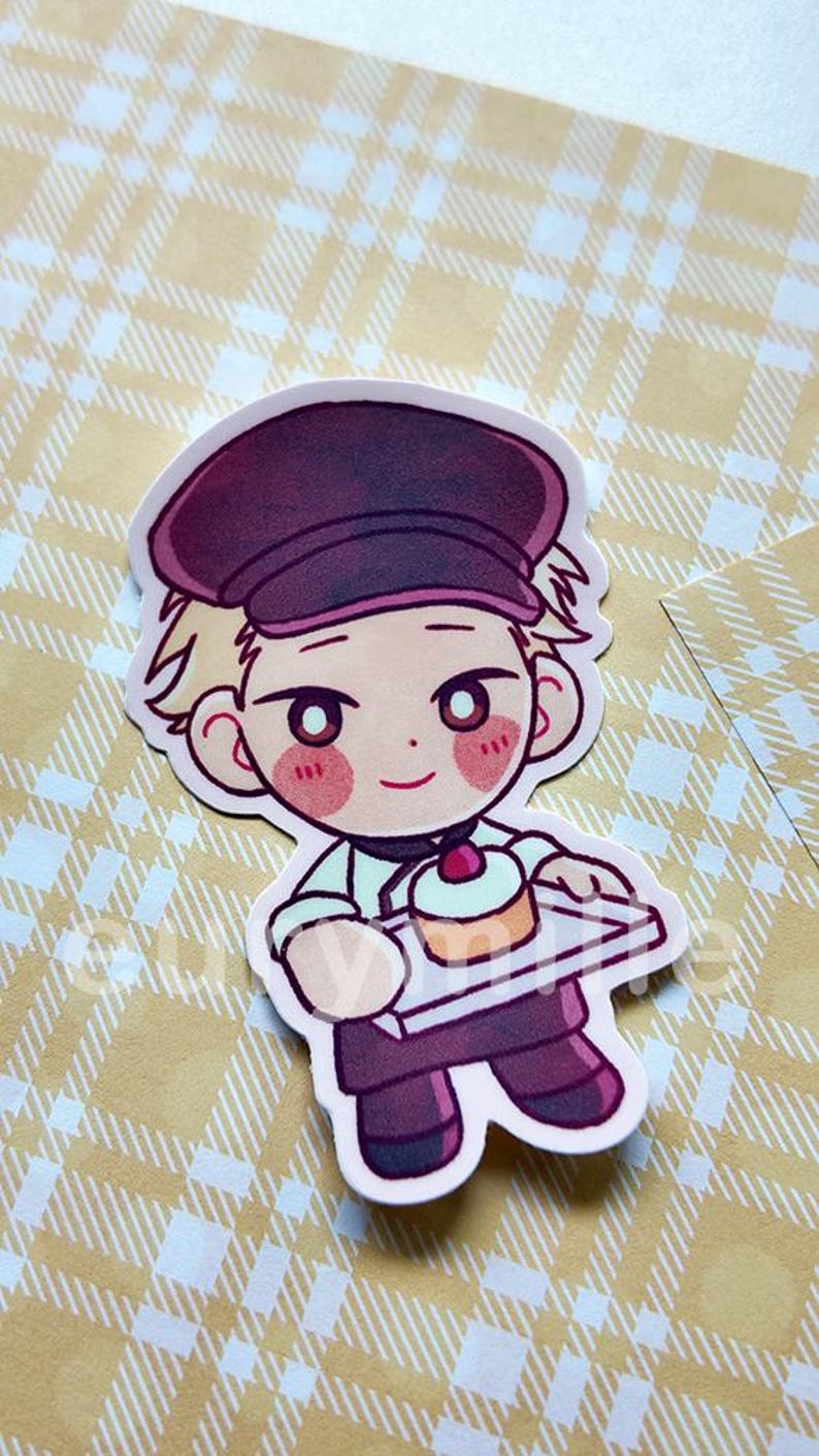 May include: A die-cut sticker featuring a cartoon character in a baker's uniform, holding a tray with a cupcake. The character has blonde hair, a maroon hat, and rosy cheeks. The background is a yellow plaid pattern.