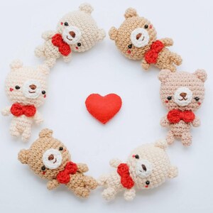 Peut inclure: Six ours en peluche crochetés, beiges et bruns, disposés en cercle, chacun avec un nœud papillon rouge. Un cœur rouge est au centre. Les ours ont des yeux noirs et des nez bruns. Le fond est blanc.