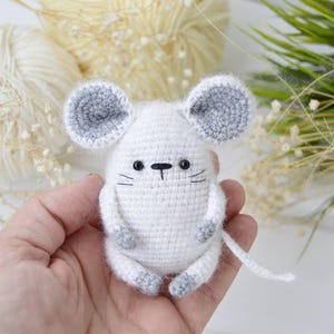 Könnte beinhalten: Eine handgefertigte weiße Häkelmaus mit grauen Akzenten an Ohren und Füßen. Die Maus hat schwarze Augen und Schnurrhaare sowie einen langen weißen Schwanz. Das Amigurumi ist ein niedliches, weiches Spielzeug.