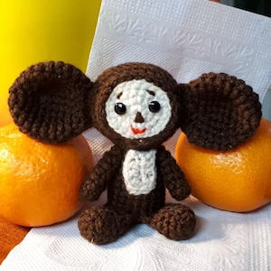 Peut inclure: Une poupée Cheburashka au crochet marron et blanc, avec de grandes oreilles, des yeux noirs et une bouche rouge. La poupée est placée entre deux oranges, sur une surface blanche.