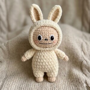 Peut inclure: Poupée au crochet faite à la main en forme de lapin. La poupée porte une combinaison de lapin crème avec une capuche et a un visage souriant avec des yeux noirs et des joues roses. La poupée est debout.