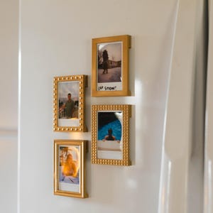 Puede incluir: Cuatro fotografías enmarcadas en oro están montadas sobre una superficie blanca. Los marcos tienen bordes decorativos y contienen varias imágenes, incluyendo una persona, una escena de piscina y un paisaje invernal. Una foto tiene el texto "1st snow".