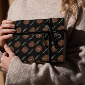 May include: A gift box wrapped in black paper with a brown and green pine cone pattern. The box is tied with a black velvet ribbon.